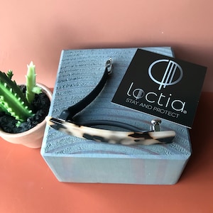 May include: A hair barrette featuring a curved, tortoiseshell-patterned acrylic top piece with a black metal clasp mechanism. Next to the barrette is a black square card with the white text "loctia" and "STAY AND PROTECT" printed below a circular logo.