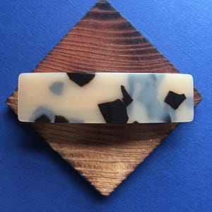 May include: Rectangular hair barrette with a cream-colored base and black and gray speckled design. The barrette is set on a wooden, diamond-shaped base with a natural wood grain pattern. The background is a solid blue.