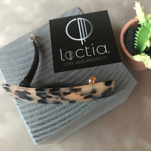 May include: A tortoiseshell hair clip with gold-colored accents and a black curved arm. The clip is resting on a gray, textured surface next to a black card with the brand name "Loctia" and the tagline "Stay and Protect."