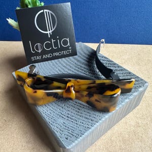 Peut inclure: Une barrette à cheveux en forme de nœud, avec un motif écaille de tortue et une bande noire. La barrette est présentée sur une surface grise texturée, avec une carte noire qui dit "Loctia STAY AND PROTECT".