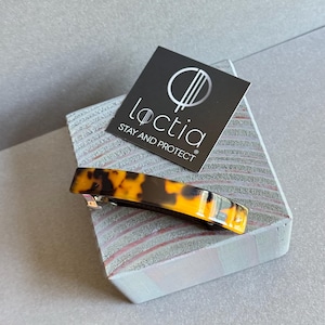 May include: A rectangular hair barrette with a tortoiseshell pattern in shades of brown and yellow. The barrette is made of a glossy material and has a silver-colored clasp. A black card with the brand name "Loctia" is also visible.
