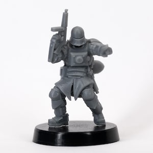 May include: A grey miniature figurine of a soldier in full armour, holding a rifle and shield. The figure is standing on a black circular base, with details including a helmet, shoulder pads, and knee guards. The figure is in a dynamic pose.