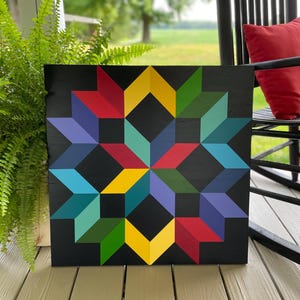 May include: A colorful geometric quilt block pattern painted on a black wooden square. The pattern features a star shape with alternating colors of red, yellow, green, blue, and teal.