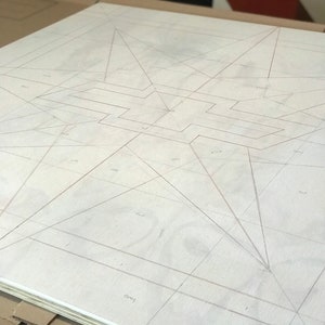May include: A white canvas with a grid pattern drawn in pencil. The grid is divided into squares and triangles. The canvas is lying on a brown cardboard box.