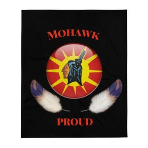 Puede incluir: Manta negra con un emblema central que representa un perfil nativo americano, un diseño de sol rojo y amarillo, y la palabra "MOHAWK" arriba. La palabra "PROUD" está debajo del emblema en rojo. Dos plumas blancas con puntas azules a cada lado.
