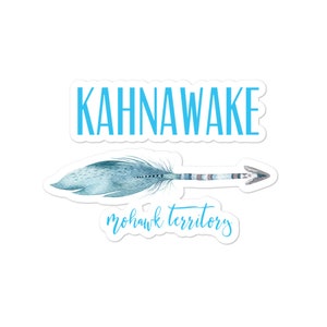 May include: A sticker with the word "KAHNAWAKE" in light blue block letters above a watercolour-style feather arrow. Below the arrow, the words "mohawk territory" are written in a script font, also in light blue.