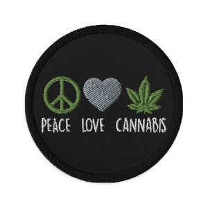 Op de afbeelding: Geborduurde zwarte patch met de woorden "PEACE LOVE CANNABIS" onder een groen vredesteken, een grijs hart en een groen cannabisblad. De patch heeft een zwarte rand.
