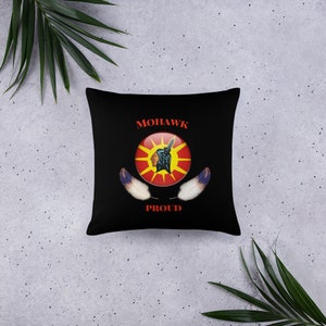 May include: Black square cushion with the words "MOHAWK PROUD" in red. The cushion features a circular emblem with a Native American profile, a raised fist, and a sunburst design in red, yellow, and black. Two white and blue feathers are below the emblem.