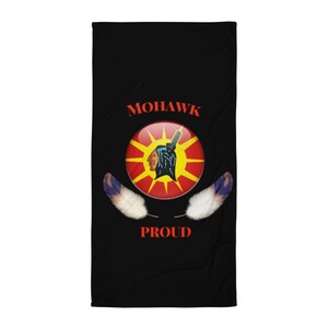 Op de afbeelding: Zwarte rechthoekige strandlaken met het woord "MOHAWK" in rood bovenaan en "PROUD" onderaan. Een cirkelvormig embleem in het midden toont een Indiaans profiel, een zwart silhouet en een zonnesterontwerp. Twee veren bevinden zich aan weerszijden.