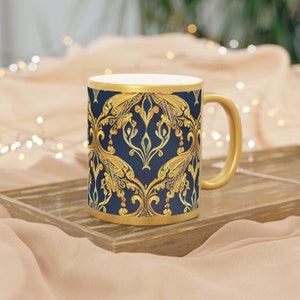 Peut inclure: Mug doré avec un fond bleu marine et un motif floral doré orné. La tasse a une anse et un bord dorés. La tasse est posée sur un plateau en bois.