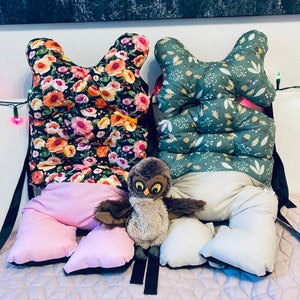 May include: A collection of decorative pillows and a stuffed owl toy. Two butterfly-shaped pillows feature floral patterns in black and green. Additional pillows are pink and off-white. The owl has brown and tan fur with large eyes.