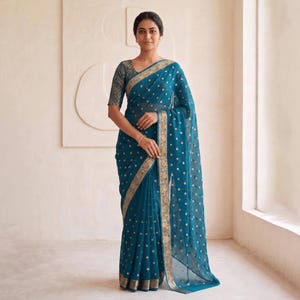 May include: A teal saree with gold polka dots and a gold border. The saree is draped over a person. The sleeves of the blouse are a darker teal with a gold pattern. The background is a light beige.