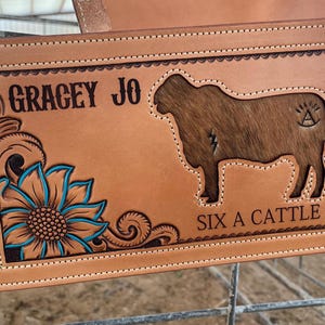May include: A brown leather saddle blanket with a sunflower design, a cow silhouette, and the text "GRACEY JO SIX A CATTLE".