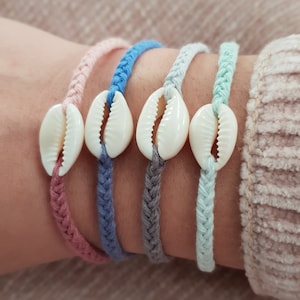 Puede incluir: Cuatro pulseras trenzadas con un dije de concha blanca en cada una. Las pulseras son rosa, azul, gris y verde claro.