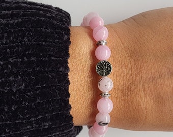 May include: Pink beaded bracelet with a silver tree of life charm. The bracelet features round, light pink beads and silver spacers. The bracelet is worn on a wrist, with a black sleeve in the background.