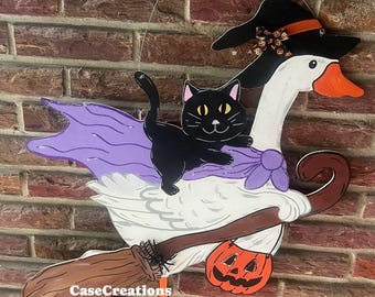 May include: A wooden Halloween decoration featuring a white goose wearing a witch hat and carrying a pumpkin. A black cat rides on the goose's back, with a purple cape. The goose holds a broom. The text "CaseCreations Shop42" is visible.