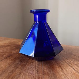 May include: A cobalt blue glass vase with a geometric, multi-faceted diamond shape. The vessel features a wide, angular body that tapers upwards into a narrow, cylindrical neck with a smooth, rounded rim, crafted from translucent blue glass.