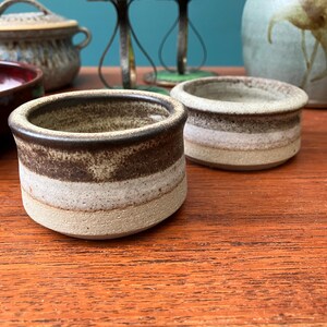 May include: Two small, handmade ceramic pots with a layered design. The pots have a dark brown rim, a light brown middle section, and a beige base. The pots are on a wooden surface.