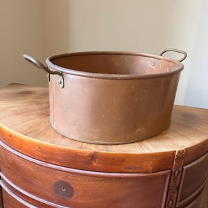 May include: An oval-shaped copper basin featuring a smooth metallic surface, a reinforced rim, and two curved metal handles attached to the sides with rivets. The basin has a warm, reddish-brown tone and a deep, open interior for storage or display.