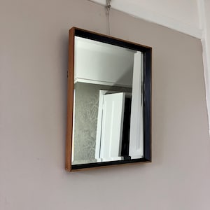 May include: A rectangular mirror with a wooden frame and a black border. The mirror is suspended on a wall. The frame is light brown. The mirror reflects a white door and a patterned wall.