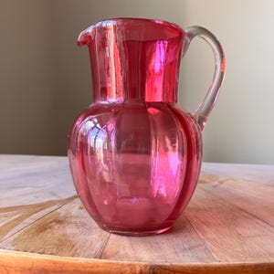 May include: A vibrant, translucent hot pink glass jug featuring a rounded, bulbous body with vertical ribbed detailing, a wide pouring spout, and a clear, curved glass handle attached to the side of the vessel for easy gripping and pouring.