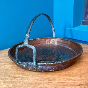 May include: An antique copper tray with a handle. The tray features a circular pattern on the base and a curved handle. The copper has a patina, indicating age and use. The tray is on a wooden surface.