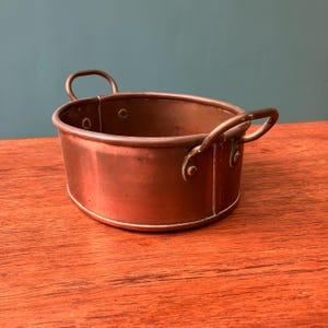 May include: A small, copper-coloured oval pot with two loop handles. The pot has a rounded base and a slightly flared rim. The copper has a warm, reddish-brown hue, and the pot appears to be vintage or antique.