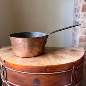 May include: A round copper saucepan with a weathered, metallic finish and a long, dark, textured iron handle attached to the side. The interior of the pan shows a silver-toned lining, and the exterior surface displays natural patina and marks.