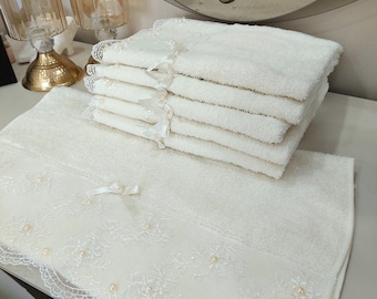May include: A stack of white bath towels with lace trim and pearl embellishments. The towels are folded and arranged on a white surface.
