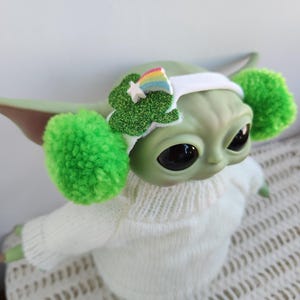 Op de afbeelding: Een groen pluche speelgoed met grote oren, met een wit hoofdband met een groen klavertjevier en een regenboogontwerp. Het speelgoed draagt een witte trui en heeft groene pompoenen aan de oren.