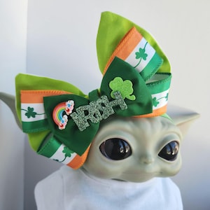 Puede incluir: Lazo de cinta verde, naranja y blanco con un trébol y "IRISH" en letras de purpurina. El lazo está sobre una figura verde y gris.