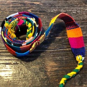 Puede incluir: Un cinturón tejido de colores con una borla trenzada. El cinturón está hecho de múltiples tiras de tela en varios colores, incluyendo rojo, naranja, amarillo, verde, azul y morado. El cinturón está enrollado y la borla cuelga.