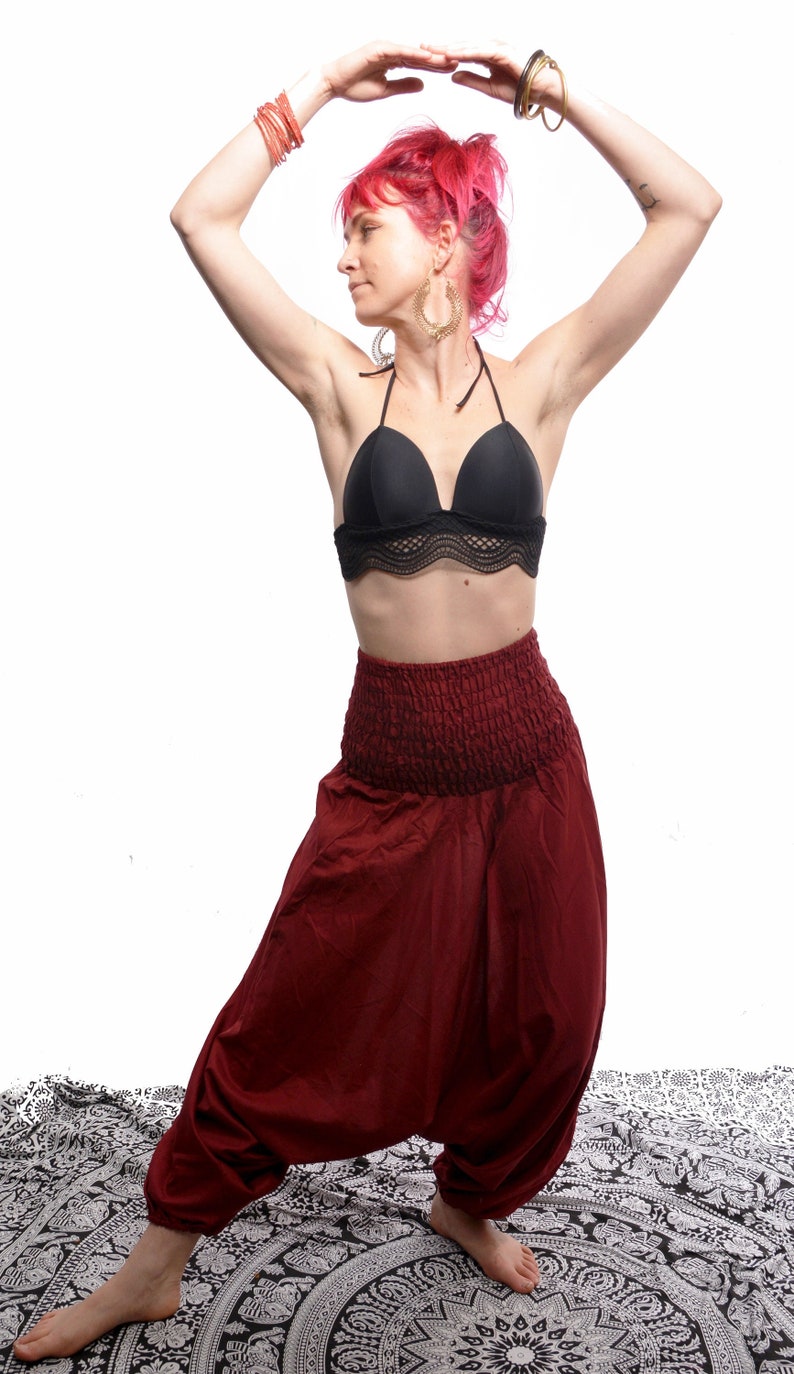 May include: A woman with pink hair wearing a black halter top and maroon harem pants. She is standing on a white and black patterned rug.