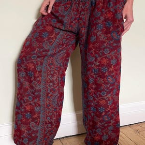 May include: Red patterned pants with a floral and paisley design. The pants have a wide leg and an elastic waistband. The fabric is a deep red with blue and gold accents. The pants are a comfortable, casual style.