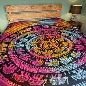 May include: A colorful bedspread with a mandala design featuring elephants. The bedspread has a black background with pink, yellow, and blue accents. The headboard is made of light-colored wood.