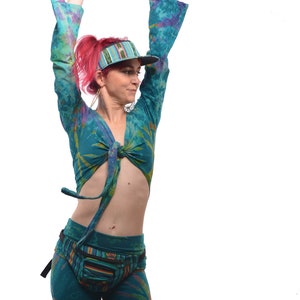 May include: A woman wearing a teal tie-dye crop top with a matching pair of pants. She is wearing a striped visor and has pink hair. She is holding her arms up in the air.