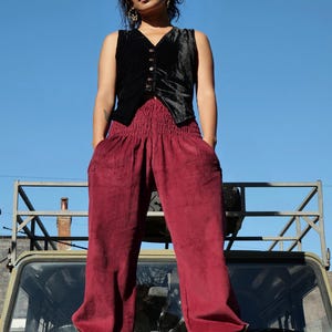 May include: A black velvet vest with a V-neckline and front buttons paired with deep red corduroy trousers featuring a wide, elasticized smocked waistband and loose-fitting legs, worn with black lace-up boots.