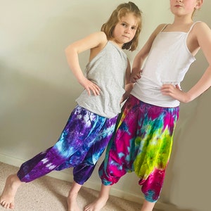 May include: Two children wearing colorful tie-dye pants. The girl on the left is wearing a gray tank top and the girl on the right is wearing a white tank top. Both girls are standing with their hands on their hips.
