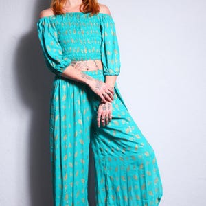 Op de afbeelding: Een turquoise off-shoulder crop top en wijde broek set met een gouden bladprint. De top heeft een gerimpelde buste en de broek heeft een wijde, vloeiende silhouet.