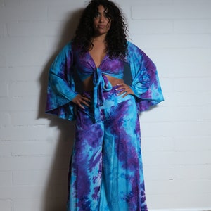 Könnte beinhalten: Ein zweiteiliges Batik-Outfit in Blau- und Lilatönen. Das Oberteil ist ein Crop-Top mit langen, ausgestellten Ärmeln und einem Bindverschluss. Die passende Hose ist weit geschnitten. Das Outfit ist aus fließendem Stoff gefertigt.