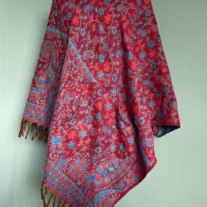 Könnte beinhalten: Ein rotes Poncho mit blauer Blumenstickerei und Kapuze. Das Poncho hat einen Fransenbesatz und wird auf einer Schaufensterpuppe präsentiert. Das aufwendige Design zeigt Paisley- und Blumenmuster.