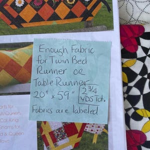 May include: A fabric sample for a twin bed runner or table runner. The fabric has a geometric design with orange, black, and white squares, and floral accents. The dimensions are approximately 51 cm x 150 cm.