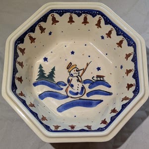 May include: Octagonal ceramic bowl with a white background and blue and brown trim. The bowl features a snowman with a blue hat and scarf, a sled, and two pine trees. The rim of the bowl is decorated with a repeating pattern of brown bells.