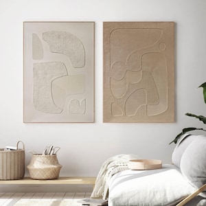 Könnte beinhalten: Zwei gerahmte abstrakte Kunstdrucke mit einem strukturierten, erhabenen Design in Beige- und Weißtönen. Die Drucke hängen an einer weißen Wand über einem hellen Sofa mit einer Wolldecke.