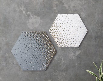 May include: Two hexagonal wall art pieces featuring a textured gradient of small, raised gold-toned metallic elements that transition across a solid gray surface on the left and a solid white surface on the right.