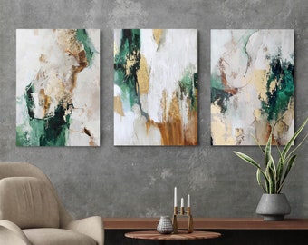 May include: Three abstract paintings in a set. The paintings are a mix of white, green, gold, and brown. The paintings are hung on a gray wall above a wooden table.