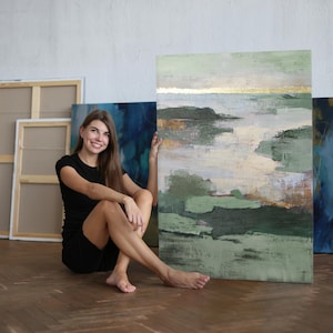May include: A woman sits on a wooden floor, leaning against a large abstract painting with green, blue, and gold hues. The painting has a textured surface and a contemporary style.