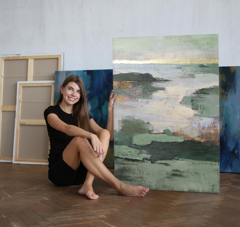 May include: A woman sits on a wooden floor, leaning against a large abstract painting with green, blue, and gold hues. The painting has a textured surface and a contemporary style.