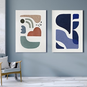 May include: Two rectangular wall art pieces featuring textured abstract shapes. The left canvas displays beige, sage green, terracotta, and dark gray forms. The right canvas features layered navy and periwinkle blue shapes on a white background.