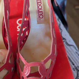 May include: Pair of pink Bandolino open-toe slingback heels. The shoes feature a cut-out design on the upper and a small heel. The brand name "Bandolino" is visible on the insole. The shoes are on a red surface.